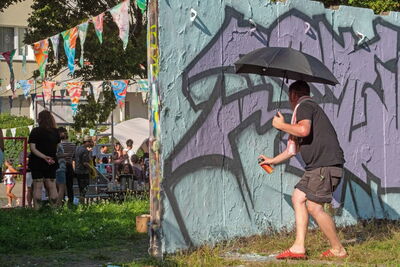 Ein Mensch schützt sich mit einem Schirm vor Sonne und sprüht mit einer Farbdose eine Wand an.