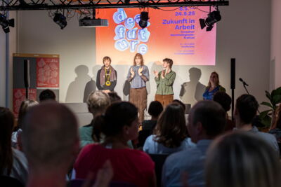 Auf der Bühne des Dear Future Festival stehen drei Menschen vor Publikum und applaudieren.